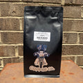 A black bag of Coffee Co. Mexico Medium Roast whole bean coffee stands upright on a rusty metal surface against a brown brick wall background.