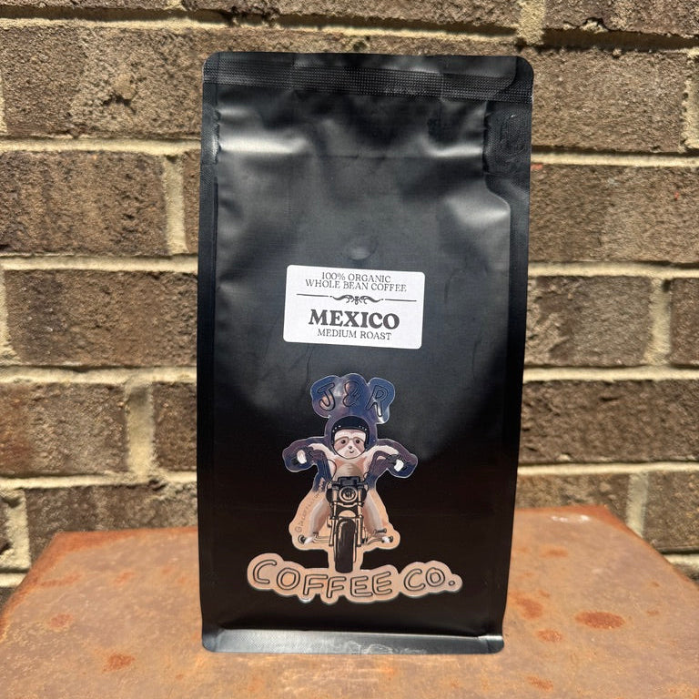 A black bag of Coffee Co. Mexico Medium Roast whole bean coffee stands upright on a rusty metal surface against a brown brick wall background.