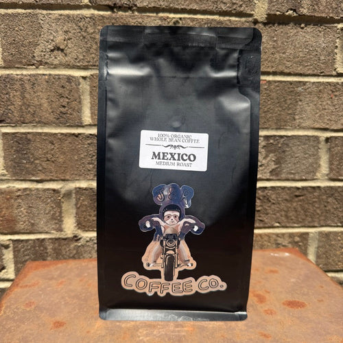 A black bag of Coffee Co. Mexico Medium Roast whole bean coffee stands upright on a rusty metal surface against a brown brick wall background.