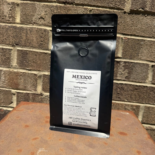 A black bag of coffee labeled 'MEXICO' stands upright on a rusty metal surface against a brick wall background.